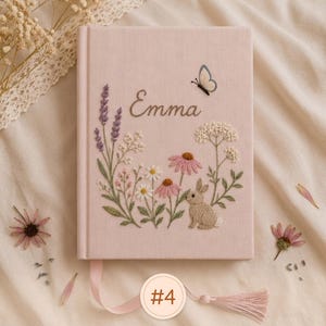 Pode incluir: Um di&aacute;rio revestido de tecido rosa claro com caules florais bordados, um pequeno coelho castanho e uma borboleta. O nome Emma est&aacute; bordado em letra cursiva sobre o design bot&acirc;nico. Um marcador de fita rosa com uma borla est&aacute; fixado.