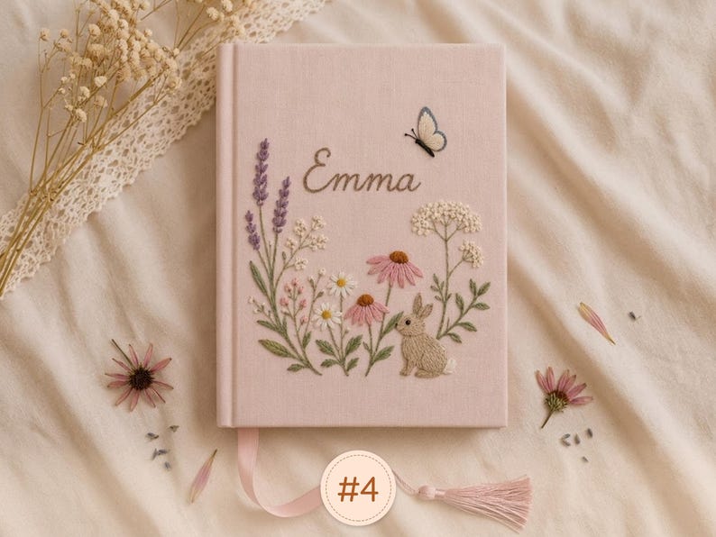 Pode incluir: Um di&aacute;rio revestido de tecido rosa claro com caules florais bordados, um pequeno coelho castanho e uma borboleta. O nome Emma est&aacute; bordado em letra cursiva sobre o design bot&acirc;nico. Um marcador de fita rosa com uma borla est&aacute; fixado.