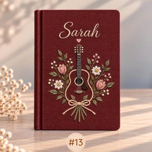 May include: A deep red fabric-covered journal featuring embroidered details of a guitar surrounded by pink and white flowers, green leaves, and a small heart. The name Sarah is embroidered in white script above the guitar. A circular tag labeled #13 is visible.
