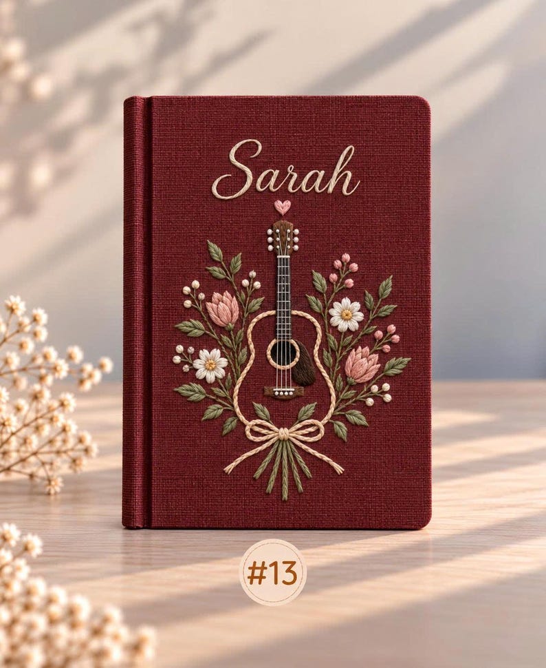 May include: A deep red fabric-covered journal featuring embroidered details of a guitar surrounded by pink and white flowers, green leaves, and a small heart. The name Sarah is embroidered in white script above the guitar. A circular tag labeled #13 is visible.