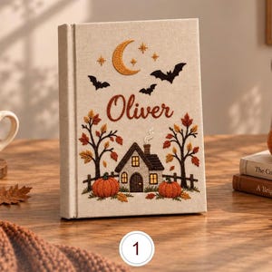 Peut inclure: Un livre recouvert de tissu beige avec des motifs brodés sur le thème de l'automne, incluant un croissant de lune, des étoiles, des chauves-souris, des arbres, des citrouilles et une petite maison. Le prénom Oliver est brodé en brun.