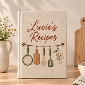 May include: A cream-coloured fabric-covered recipe book with the embroidered text "Lucie's Recipes" in rust-coloured thread. Below the text, a string holds embroidered kitchen items: a green pan, a brown cutting board, a whisk, a spatula, and a floral oven mitt.