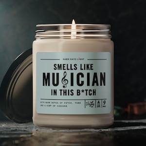 May include: A beige candle jar with a black lid. The label reads "Smells Like Musician In This B*tch" with a treble clef symbol. The candle is vegan and has a burn time of 9 hours.