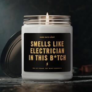 May include: A white candle jar with a black label that reads "HARD DAYS LIGHT" and "SMELLS LIKE ELECTRICIAN IN THIS B*TCH". The candle is lit and the jar has a black lid.