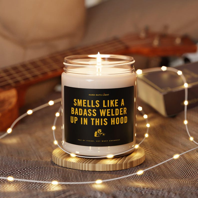 May include: A lit candle in a glass jar with a black label that reads "Smells Like a Badass Welder Up in This Hood." The candle sits on a round wooden coaster, surrounded by string lights. A guitar is visible in the background.