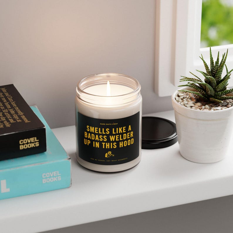 May include: A lit, white candle in a clear glass jar with a black label that reads "Smells Like a Badass Welder Up in This Hood." The candle sits on a white shelf next to a small potted succulent and a stack of books.