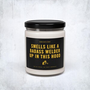 May include: A white candle in a glass jar with a black lid. The label is black with yellow text that reads "Smells Like a Badass Welder Up in This Hood." Below the text is a small graphic of a welder.