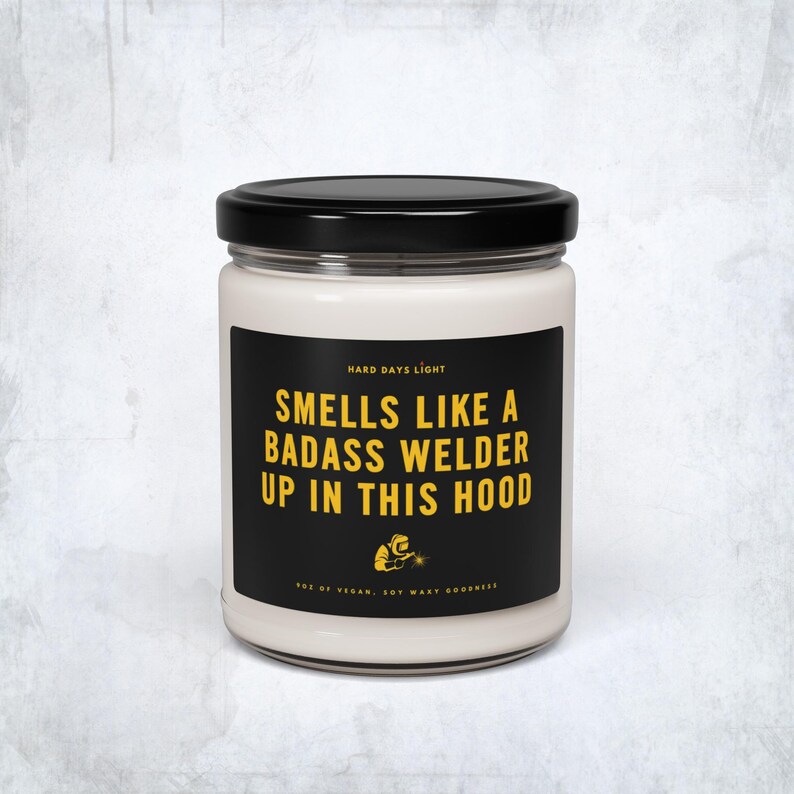 May include: A white candle in a glass jar with a black lid. The label is black with yellow text that reads "Smells Like a Badass Welder Up in This Hood." Below the text is a small graphic of a welder.