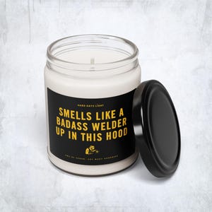 May include: A white soy wax candle in a clear glass jar with a black lid. The candle has a black label with yellow text that reads "Smells Like A Badass Welder Up In This Hood."