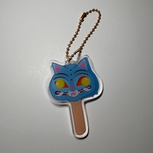 以下が含まれることがあります： 木製の棒が付いた青い猫の顔のアイスキャンディーの形をした透明なアクリルキーホルダー。猫のデザインには、大きな黄色い目、ピンクの耳、歯を見せた笑顔が描かれています。上部には金色の金属製ボールチェーンが付いています。