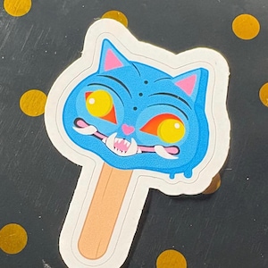 以下が含まれることがあります： 木製の棒が付いた青い猫の顔のアイスキャンディーの形をしたダイカットステッカー。猫は明るい黄色の目、ピンクの耳の内側、小さなピンクの鼻、鋭い白い牙を見せる広い口を持っています。デザインは太い輪郭が特徴です。