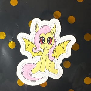 May include: A die-cut sticker featuring a yellow pony character with pink hair, large red eyes, and small fangs. The character has yellow bat-like wings and a pink tail. The design includes a white border and a small signature near the bottom edge.