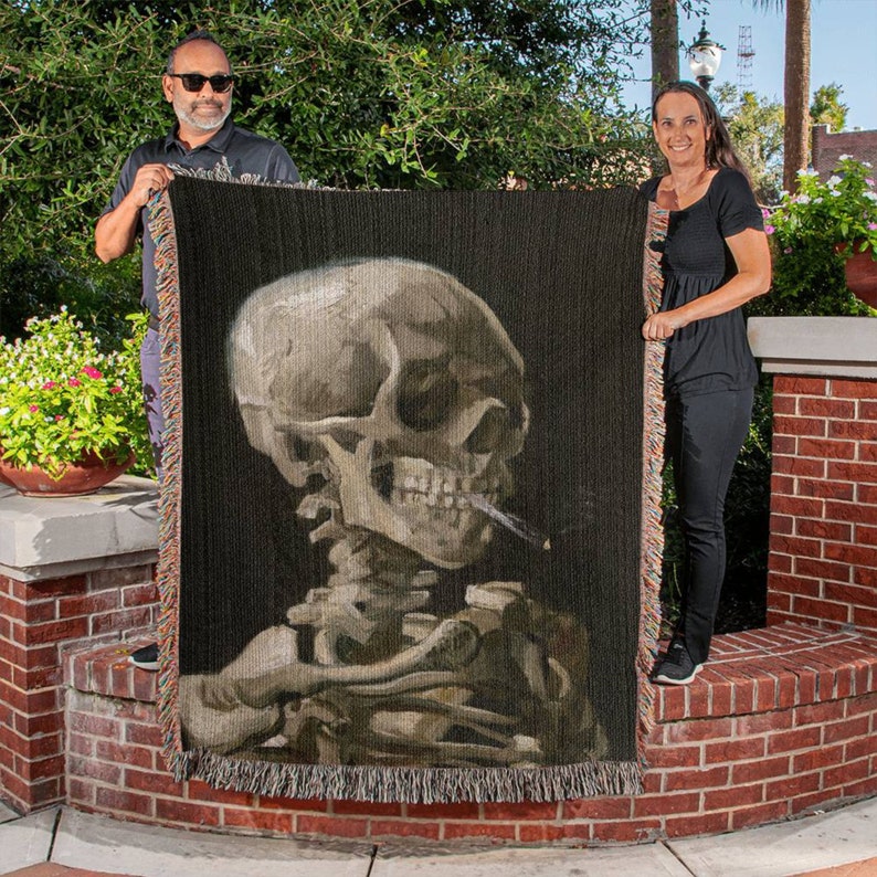 May include: A woven tapestry wall hanging featuring a detailed depiction of a human skull with a cigarette in its mouth. The skull is rendered in shades of gray and white against a dark background.