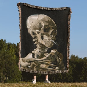 May include: A woven tapestry throw blanket featuring a painting of a human skull with a cigarette in its mouth. The skull is depicted in a realistic style with detailed bone structure and a dark background.