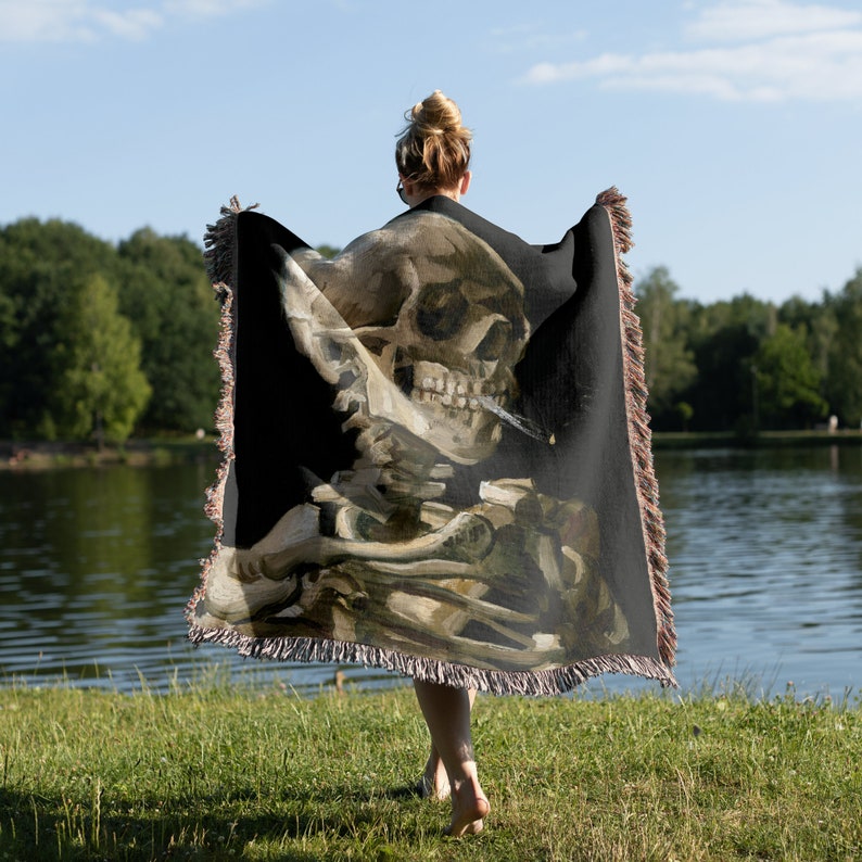 May include: A black woven throw blanket with a printed image of a skull holding a cigarette. The skull is depicted in a realistic style with a detailed bone structure and a cigarette in its mouth. The blanket is fringed on all sides.