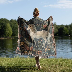 May include: A woman holding a tapestry throw blanket with a unicorn and floral design. The blanket is dark blue with gold, red, and white accents.