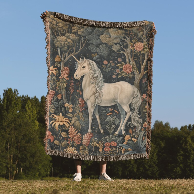 May include: A woven tapestry throw blanket features a white unicorn standing in a lush forest with colorful flowers and leaves. The tapestry has a fringe border.
