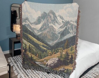 May include: A woven tapestry throw blanket featuring a scenic mountain landscape with a river flowing through a valley. The mountains are depicted in shades of blue, green, and brown, with snow-capped peaks. The throw blanket has a fringed border.
