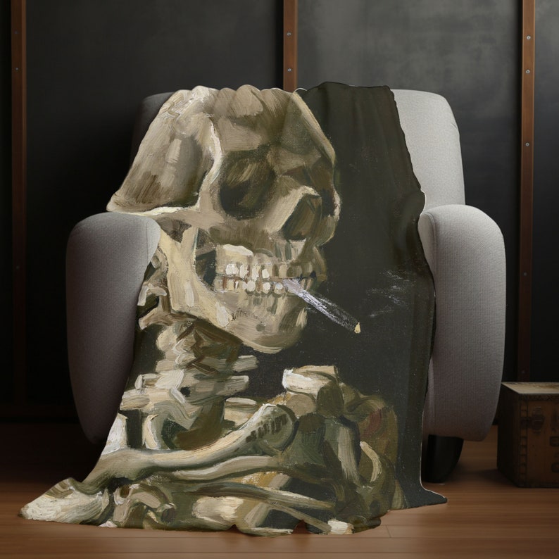 May include: A throw blanket featuring a painting of a human skull with a cigarette in its mouth. The skull is painted in a realistic style with visible teeth and a dark background.