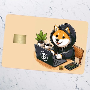 Pode incluir: Um cão Shiba Inu vestindo um moletom com capuz preto está sentado em uma mesa com um laptop, uma xícara de café e uma planta. O laptop tem um símbolo de Bitcoin na tela. O cão está digitando no teclado.