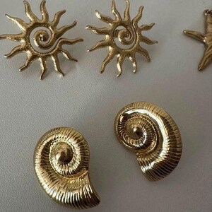 May include: Gold-coloured earrings in various shapes, including sunbursts, seashells, and a starfish. The earrings have a shiny, metallic finish and are arranged on a light-coloured surface. The sunburst earrings have a spiral centre.