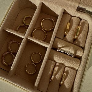 May include: A beige jewellery case displaying an assortment of gold earrings and a ring. The earrings include small hoops, hexagonal shapes, and drop styles. The ring is a delicate band with small, clear stones. The case is open, revealing the organised compartments.