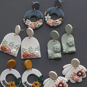 Könnte beinhalten: Eine Sammlung handgefertigter Polymer-Ton-Ohrringe in verschiedenen Formen und Farben. Designs beinhalten florale und botanische Elemente, mit Farbtönen von Blau, Weiß, Grün und Gelb. Einige Ohrringe sind kreisförmig, andere bogenförmig oder fächerförmig, alle mit goldfarbenen Stiften.