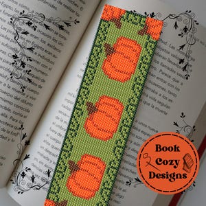 Puede incluir: Un marcapáginas de cuentas con un fondo verde y calabazas naranjas. El marcapáginas tiene un borde decorativo y las palabras "Book Cozy Designs" en un círculo naranja.