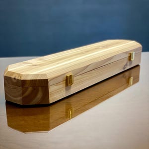 May include: A rectangular wooden box with a light tan natural grain finish and beveled edges. The box features two gold-colored metal latches on the front side, securing the lid to the base. The wood displays visible knots and linear grain patterns.