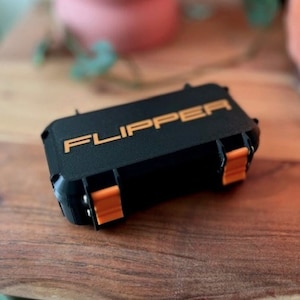 May include: Black rectangular case with the word "FLIPPER" in orange lettering. The case has orange clasps and hinges. The case is on a wooden surface.