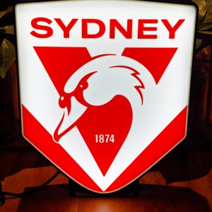 May include: A rectangular illuminated sign featuring the word "SYDNEY" in red, above a white swan emblem on a red and white shield. The number "1874" is below the swan. The sign is on a wooden surface.