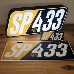 May include: A black, yellow, and white sign displaying "SP 433 #10". A matching keychain and license plate are also present. The sign is rectangular with rounded corners and a black border. The keychain has a silver ring.