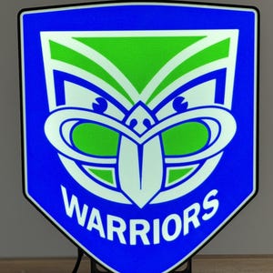 May include: A blue, green, and white Warriors logo light. The logo is a shield shape with a stylized face design. The word "WARRIORS" is written in white at the bottom of the shield. The light is on a black stand.