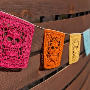 May include: A string of colourful crocheted skull-shaped flags hanging on a wooden fence. The flags are pink, orange, yellow, blue and green.