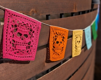 May include: A string of colorful crocheted skull-shaped flags hanging on a wooden fence. The flags are pink, orange, yellow, blue, and green.