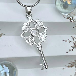 May include: A silver key pendant necklace. The pendant has a floral design with a central clear stone. The key hangs from a silver chain. The background includes decorative glass spheres and greenery.