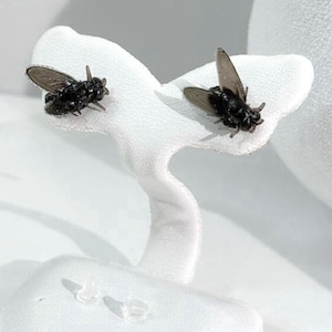 Peut inclure: Une paire de boucles d'oreilles noires en forme de mouche. Les boucles d'oreilles sont présentées sur un présentoir à bijoux blanc sculpté. Les mouches ont des ailes et des corps détaillés, et sont positionnées comme si elles étaient en vol. Le fond est blanc doux.