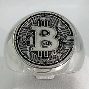 Könnte beinhalten: Ein silberner Ring mit dem Bitcoin-Symbol. Der Ring hat eine runde Oberfläche mit dem Bitcoin-Logo in der Mitte, umgeben von einem Leiterplattenmuster. Der Ring besteht aus glänzendem Silbermetall.