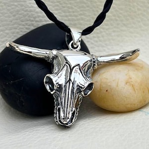 May include: A silver-colored bull skull pendant with long horns, suspended from a black cord. The pendant is highly detailed, with visible facial features. The necklace is displayed on a black and beige stone.