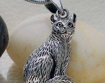 Puede incluir: Un collar con un colgante de plata en forma de gato. El gato está sentado con la cola enroscada alrededor de sus patas. El colgante está detallado con un patrón de piel texturizado.