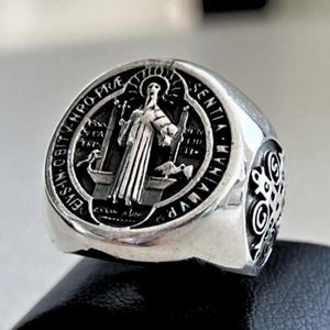 Puede incluir: Un anillo de plata con una imagen en blanco y negro de San Benito. El anillo tiene las palabras "Eius in obitu nostro" y "Sentiam vim nominis" alrededor de la imagen.