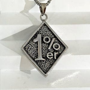 May include: Silver pendant necklace with a diamond-shaped pendant. The pendant features the text "1%er" and a textured background. The necklace is displayed against a white background. The chain is silver.
