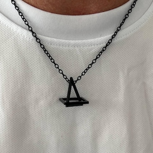 May include: Black geometric pendant necklace. The pendant features a square base with a triangle above it, suspended from a black chain. The necklace is worn against a white shirt.