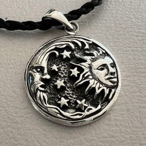 Puede incluir: Un collar con un colgante de plata que presenta un diseño detallado de un sol y una luna con estrellas. El colgante es redondo y tiene una superficie texturizada.