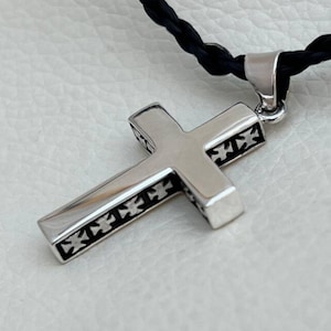 Puede incluir: Un colgante de cruz de plata con un diseño detallado de pequeñas cruces en los bordes. El colgante está unido a un cordón trenzado negro.