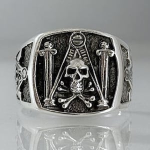 Puede incluir: Anillo plateado con un diseño de calavera y huesos cruzados, con un símbolo de compás y escuadra. El anillo tiene una superficie detallada y texturizada y una banda ancha. El diseño incluye dos espadas a los lados.
