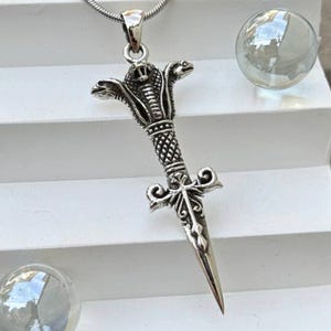 Könnte beinhalten: Eine silberfarbene Halskette mit Anhänger, die ein Dolchdesign mit einem Kobrakopf und aufwendigen Details zeigt. Der Anhänger hängt an einer feinen, strukturierten Kette. Der Dolch hat eine Spitze und einen kreuzförmigen Parier.