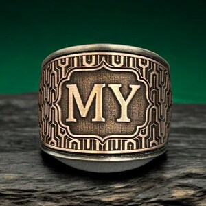 Puede incluir: Un anillo de banda ancha con acabado en bronce metálico y una placa rectangular central que muestra las letras MY en una fuente serif negrita. La superficie está decorada con un patrón geométrico repetitivo de líneas y formas entrelazadas.