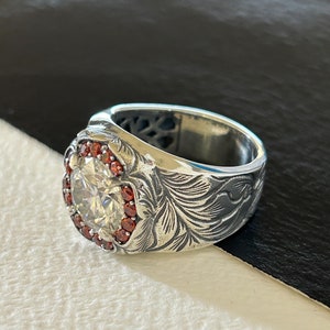 May include: A silver ring with a large, clear center stone surrounded by small red stones. The ring has an ornate floral design engraved on the band. The ring is set against a black and white background.