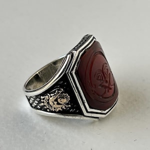 May include: A silver ring with a red carnelian stone set in a hexagonal bezel. The stone has an intricate design carved into it. The band of the ring is decorated with a gold design.